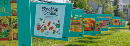 A row of large, illustrated book pages from the children’s book Rooftop Garden is displayed outdoors along a fence, creating a “storywalk” experience. The pages, mounted on teal boards, show colorful drawings of gardening scenes and tools. The grassy area