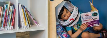 A little kid sitting in a reading nook holding book while wearing a space helmet. 