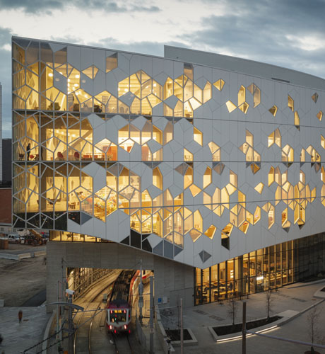 The modern Central Library with its distinctive geometric facade illuminated at dusk. A CTrain rail vehicle passes underneath the building.