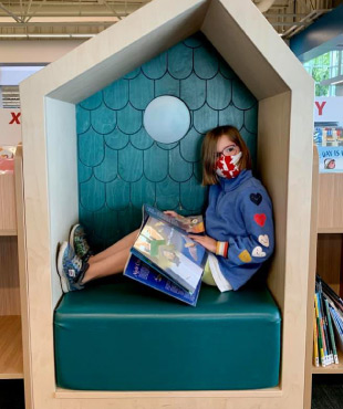 School aged girl sitting in reading nook with a book on her lap at a library.