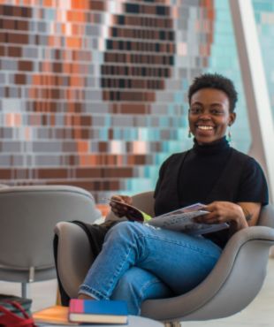 A smiling woman sits in a chair with an open book on her lap in front of a mural.