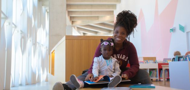 A woman with a young child sitting on her lap are looking at a picture book. They are in a colourful library space with large windows and pink walls.