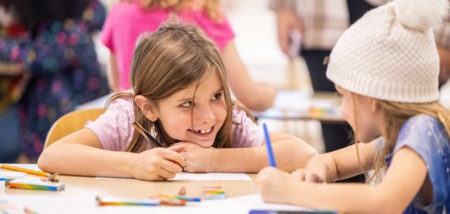 Two girls are sitting at a table colouring on pieces of paper. The background has other children doing similar crafts.