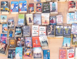 A table overflowing with a variety of paperback books, stacked and arranged in no particular order. Titles visible include "Knife," "City of Girls," "Three Women," and "The Daughter of a Black Lake."