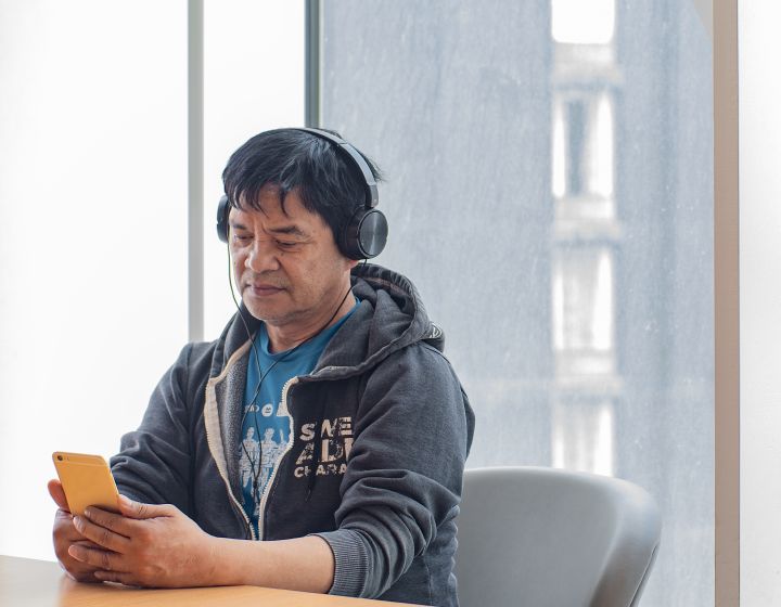 background image: A man us sitting at a table looking at his phone wearing headphones.