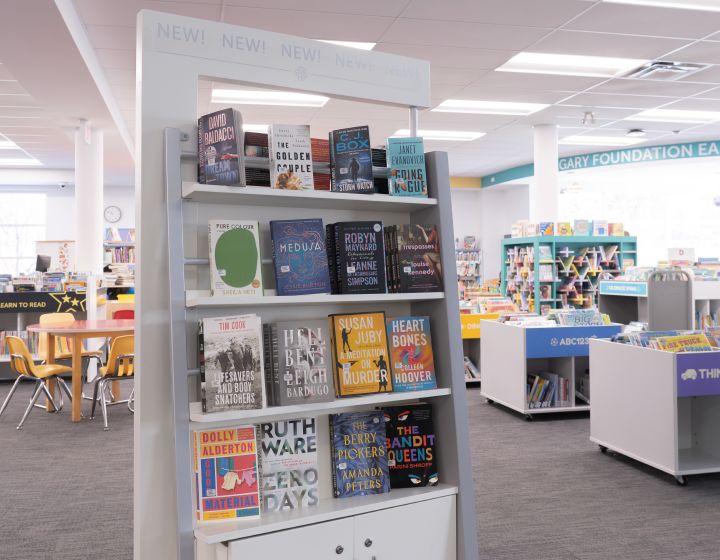 background image: A photo of a library aisle with bookshelves. Books are displayed on the shelves, with titles like "Medusa," "Heart Bones," and "Zero Days" visible.