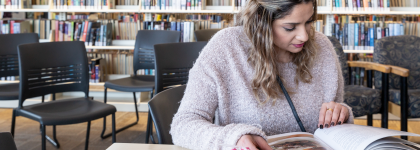 A woman with a pink sweater is sitting in a library at a table reading a book.