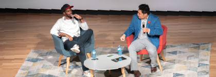 Two men sitting on chairs on a stage, engaged in a conversation. They are sitting beside a round table with water bottles. The stage has a geometric rug.