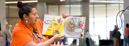 A woman wearing an orange shirt reads a colourful illustrated children's book aloud. The background shows a well-lit library.