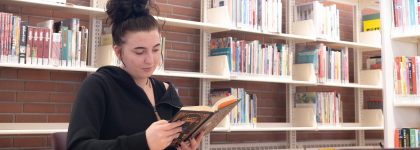 A woman with dark hair and a black sweater is sitting in a library in front of bookshelves reading a book. 