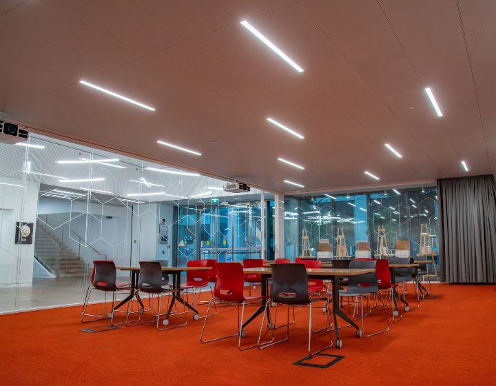 background image: This is a visual of the BMO Community room. The carpet is orange, and on top of it are tables and chairs set for four people. In the background, there are glass windows with hexagon-shaped illustrations etched in them.