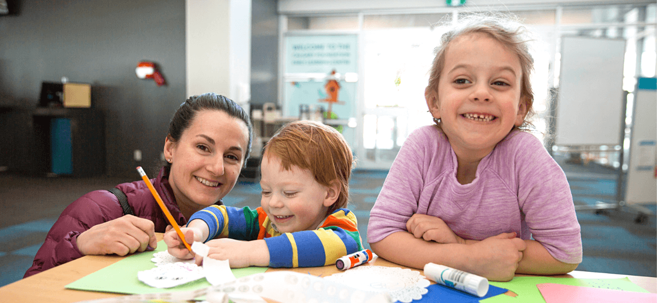 background image: A woman and two children doing a drawing activity.