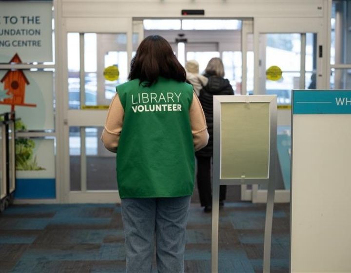 background image: A person wearing a green vest that has Library Volunteer on the back standing at the front in Crowfoot Library.