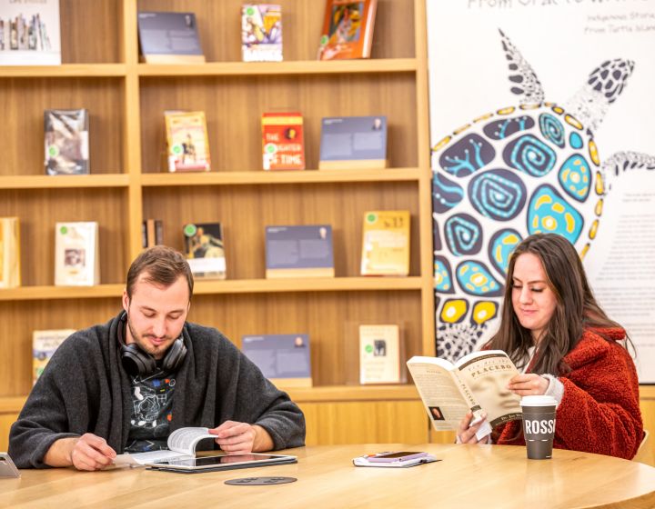 A man and a woman sitting at a table reading books. There is a bookshelf in the background as well as a mural of a turtle on the wall.