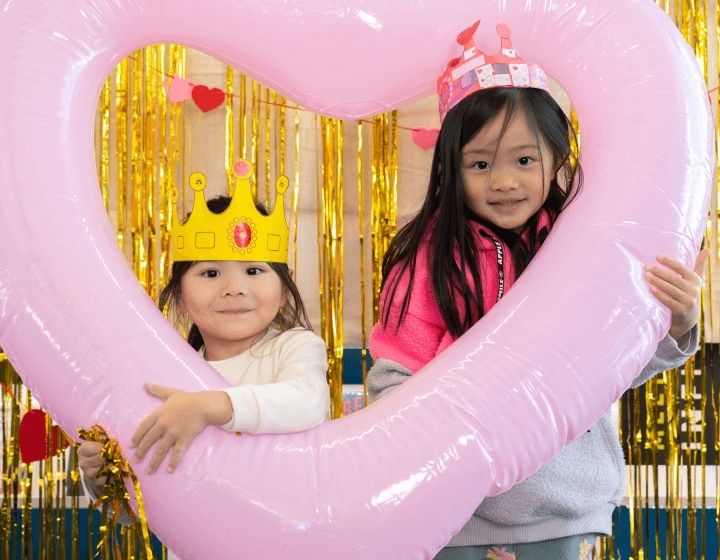 Two little girls, one on the left in a white shirt and wearing a gold crown, and the other on the right in a pink jacket and wearing a pink crown, are holding a heart balloon that frames their faces.