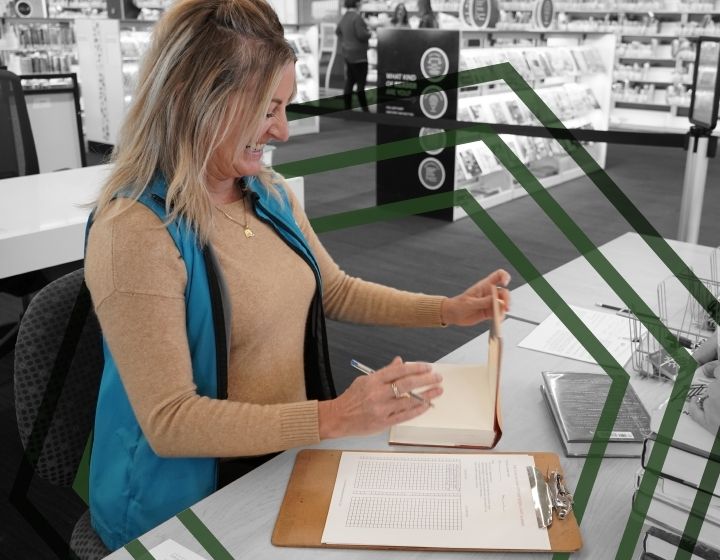 background image: A staff member sitting at a desk smiling and writing a barcode from a Library book onto a piece of paper. 